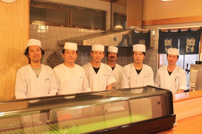 店内カウンターの中に立つ、板前帽子と白い調理衣を着た、男性従業員6人の写真