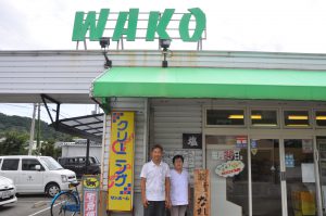屋根の上にWAKOという緑色の看板のあるお店の前に立つ、男性従業員と女性従業員の2人の写真