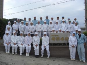 会社の看板を囲み、3列に並んだ、工場用白衣を着た、35名の従業員の写真
